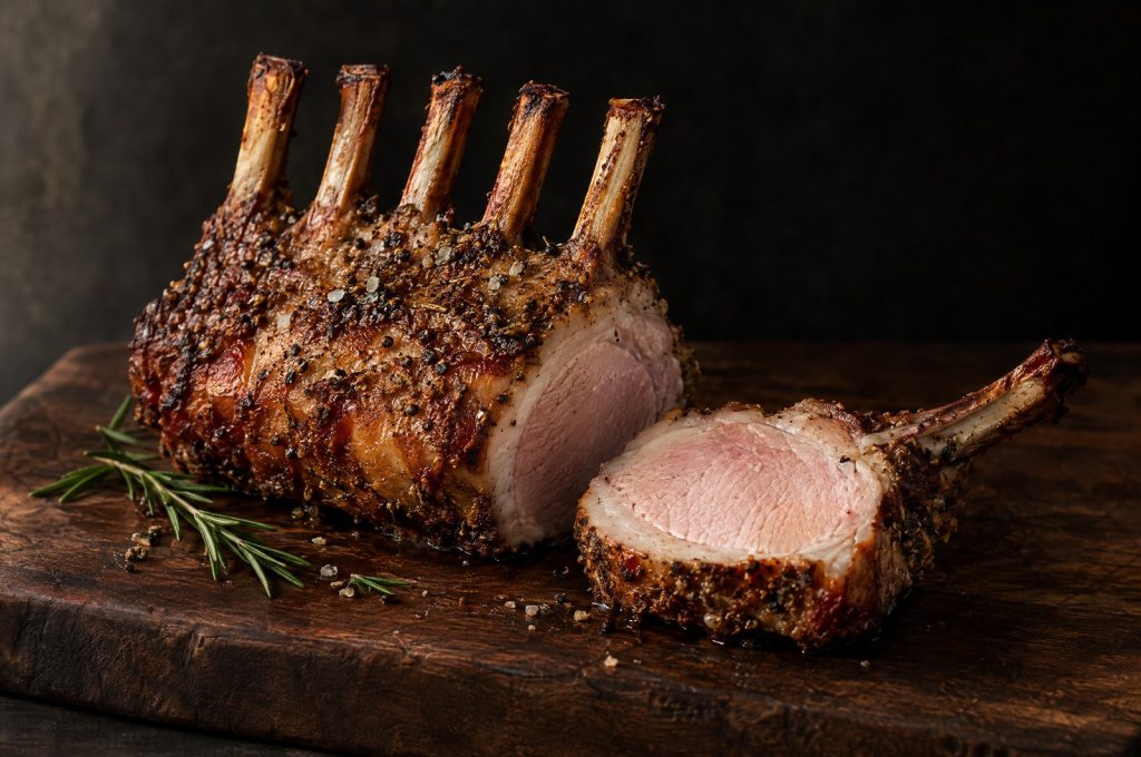 Rack of pork: a roasted 5 rib bone in pork rack with a peppered crust on a dark walnut board, one chop sliced away showing a pale pink center